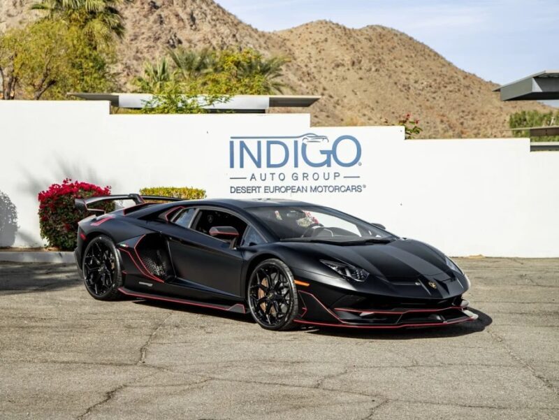 A black Lamborghini with red accents, showcasing its roaring V12 engine, is parked in front of a sign reading "INDIGO Auto Group Desert European Motorcars," set against a stunning mountainous backdrop. This exotic car is now for sale.