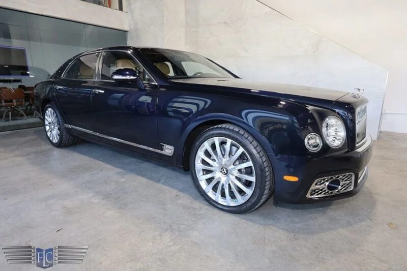 Luxury dark blue sedan, an exotic car for sale, parked indoors. Viewed from the side, it boasts silver alloy wheels and a logo in the bottom left corner of the image.