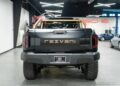 Rear view of a black Rezvani Hercules 6x6 truck parked indoors, showcasing bold orange lettering and rugged design elements, now available for sale.