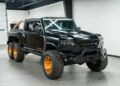 The black Rezvani Hercules 6x6 truck, featuring large off-road tires and gold rims, is parked indoors with a rugged design and a visible roll bar at the back. This impressive vehicle is now for sale.