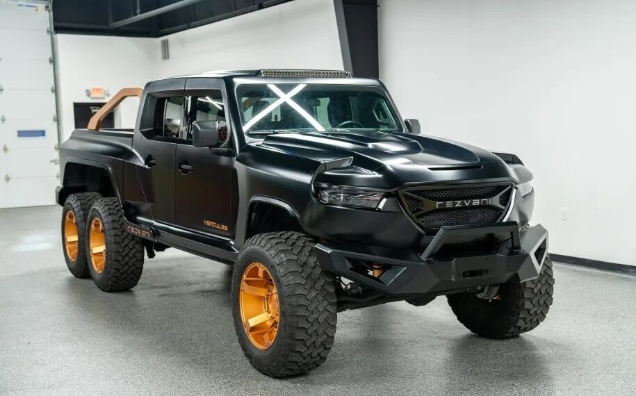 The black Rezvani Hercules 6x6 truck, featuring large off-road tires and gold rims, is parked indoors with a rugged design and a visible roll bar at the back. This impressive vehicle is now for sale.