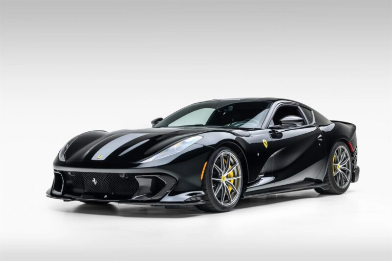 A black Ferrari 812 Competizione sports car with yellow brake calipers is parked in a white studio setting, highlighting its striking design and exclusivity.