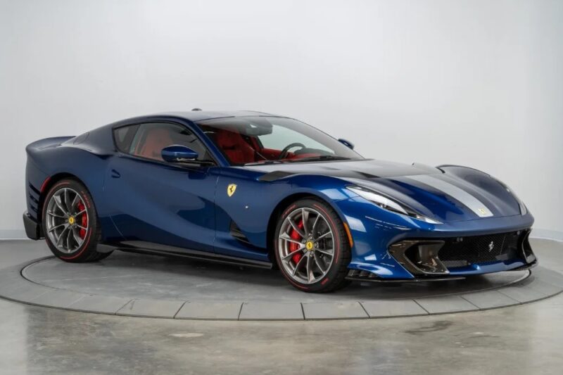 A Blu America Ferrari 812 Competizione with striking red brake calipers is parked indoors on a circular platform against a plain white background, highlighting this stunning sports car now for sale.