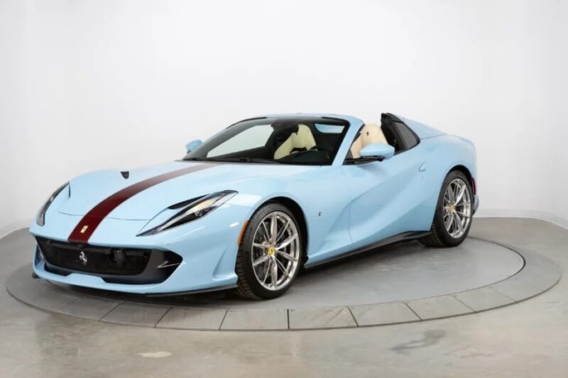 This light blue sports car, featuring a maroon stripe on the hood and silver wheels, boasts a beige interior and is magnificently displayed on a turntable in the showroom. Among exotic cars for sale, it's an eye-catching masterpiece.