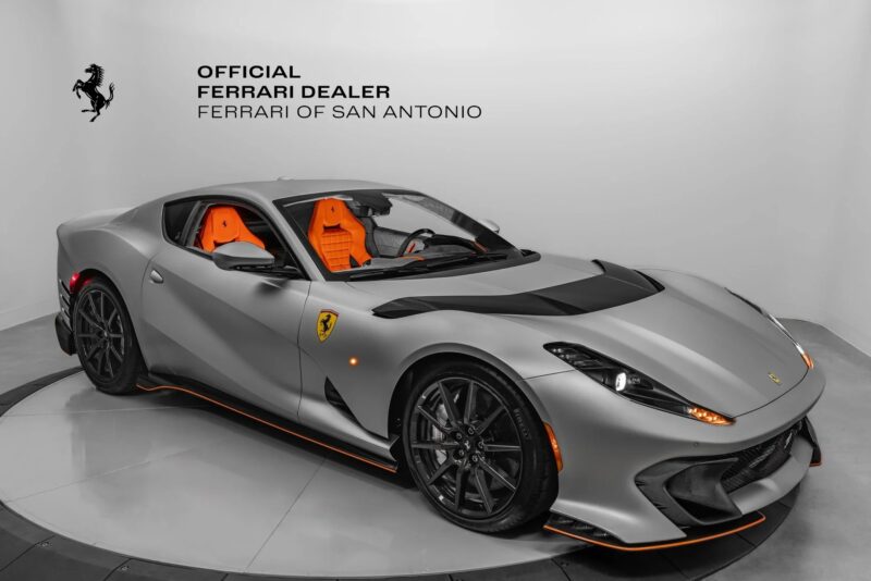 A silver Ferrari 812 Competizione with orange interior is displayed in a showroom with "Official Ferrari Dealer Ferrari of San Antonio" text and Ferrari logo on the wall, available for sale.