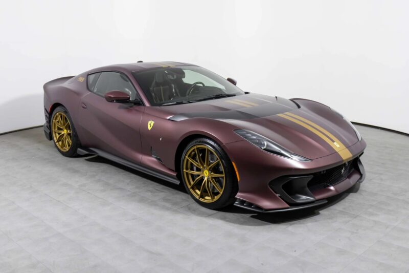 A matte maroon Ferrari 812 Competizione with gold racing stripes and wheels, for sale, parked in a plain white studio setting.