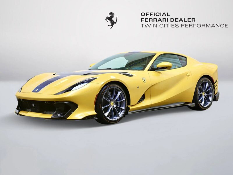 A yellow Ferrari 812 Competizione with black racing stripes is displayed in a studio setting, the Twin Cities Performance Ferrari dealer logo in the background, highlighting this stunning model now available for sale.