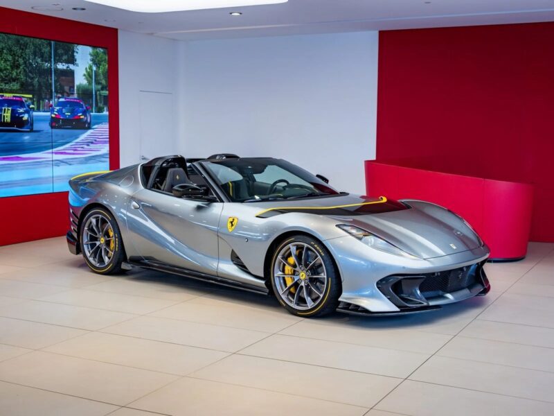 A silver Ferrari 812 Competizione convertible sports car is displayed in a modern showroom with red and white walls, a large screen showing a racing scene, and marked as for sale.