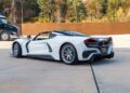 A sleek white sports car, reminiscent of the Hennessey Venom F5 Roadster, rests on a concrete surface, framed by lush trees and a large truck in the background.