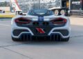 Rear view of a white sports car with blue stripes, reminiscent of the Heritage Series, parked on pavement. Two large exhausts are centered above a license plate. Trailer with branding is visible in the background, hinting at the stunning design of a Hennessey Venom F5.