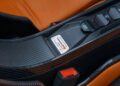 Close-up of a vehicle interior showcasing a carbon fiber panel with buttons and a label reading "Hennessey Venom F5," alongside orange leather upholstery and a visible seatbelt latch, capturing the sleek essence of this Roadster.