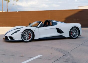 A sleek white roadster is parked on a concrete surface, with a rustic wooden fence in the background, exuding the allure of a Heritage Series masterpiece.