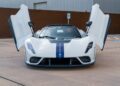 A white Heritage Series sports car, showcasing its butterfly doors and blue racing stripe, is elegantly parked in a lot near a brown wall.