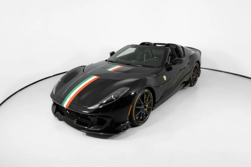 A black Ferrari 812 Competizione with an Italian flag stripe on the hood is parked in a white studio setting, showcasing its sleek design and sporty presence. This stunning vehicle is now for sale.