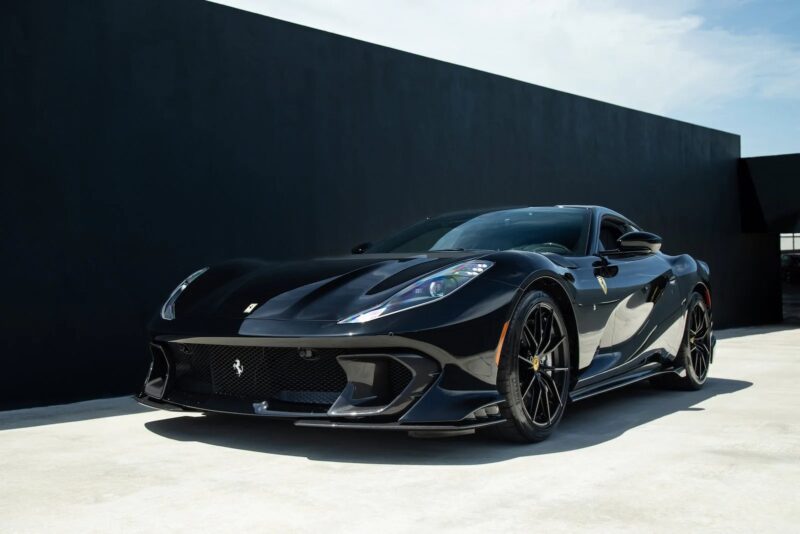 A sleek black Ferrari 812 Competizione with angular lines is parked on a concrete surface in front of a minimalist, dark wall under a partly cloudy sky—an extraordinary sports car for sale.