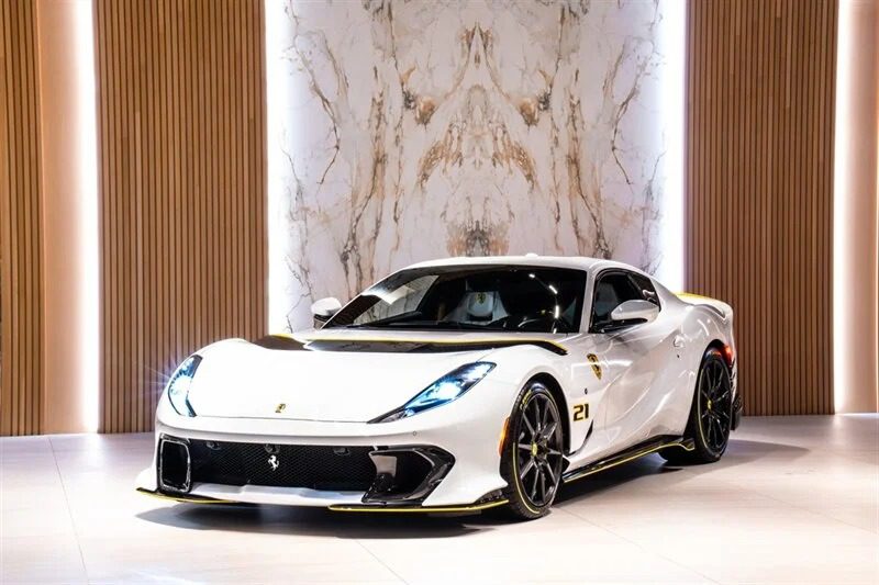 A Ferrari 812 Competizione in striking white with black and yellow accents is displayed in a modern showroom with marble and wood panel walls, now for sale.