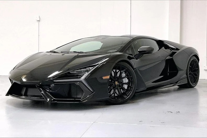 The sleek black sports car, reminiscent of the 2024 Lamborghini, features sharp design lines, a low profile, and large dark alloy wheels. It sits elegantly in a minimalist white room, capturing the essence of luxury and performance.