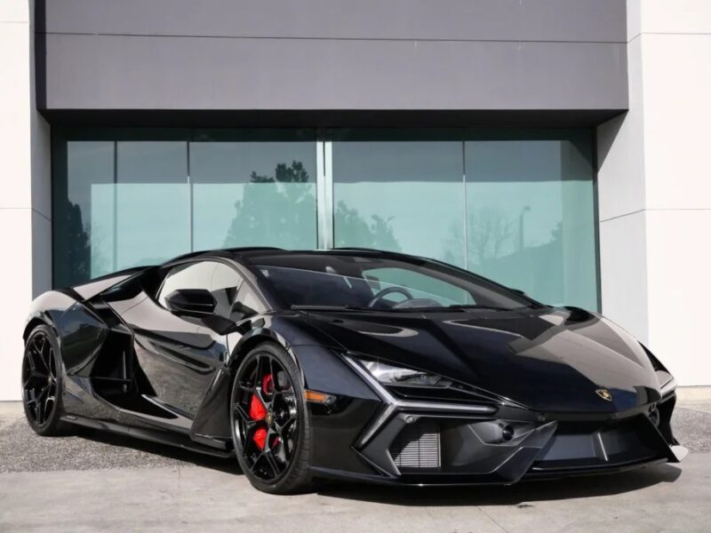 A sleek black 2024 Lamborghini sports car with red brake calipers is parked in front of a modern building with large glass panels, making it a perfect spot for someone looking to buy a Lamborghini.