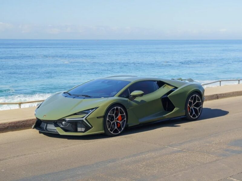 A sleek 2024 Lamborghini Revuelto sports car is parked by the ocean on a sunny day, with a clear blue sky and calm water in the background.