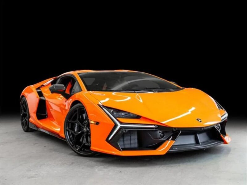 A 2024 Lamborghini Revuelto sports a bright orange hue with a sleek aerodynamic design, sharp angles, and black accents. It sits majestically parked indoors against a striking black background, inviting true enthusiasts to buy Lamborghini excellence.