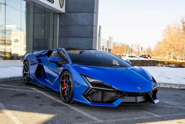 A sleek 2024 Lamborghini sports car, boasting black accents and red brake calipers, is parked outside a dealership against a snowy landscape. This Lamborghini Revuelto is an eye-catching blend of style and performance, ready for sale to those who crave luxury on wheels.