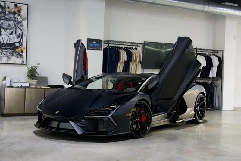 A sleek black 2024 Lamborghini Revuelto with upward-opening doors is parked indoors, exuding luxury and style. In the background, clothing hangs on a wall rack, hinting at a lifestyle synonymous with this masterpiece.