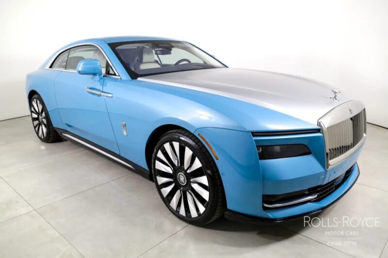 A blue and silver Rolls-Royce Spectre, an ultra-luxe electric super coupe, is parked in a showroom, showcasing its distinctive grille and large wheels.