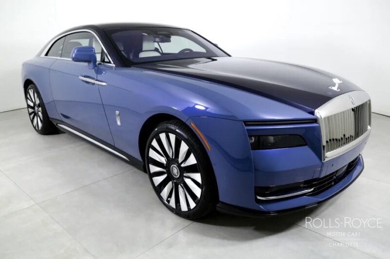 The ultra-luxe blue Rolls-Royce Spectre Electric Super Coupe gleams in a bright indoor space, effortlessly showcasing its sleek design and pristine white wall tires.