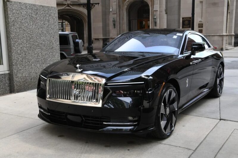A black Rolls-Royce Spectre with a sleek design is parked on an urban street, showcasing a shiny silver grille and distinctive headlights, embodying electric ultra-luxe elegance.