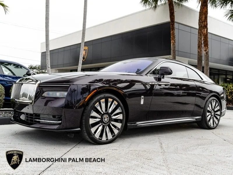 The Rolls-Royce Spectre, an ultra-luxe electric super coupe, elegantly stands on a light gray surface, juxtaposed against a modern building adorned with swaying palm trees.