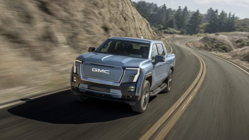 A silver GMC pickup truck drives on a winding road through a mountainous area with clear skies.