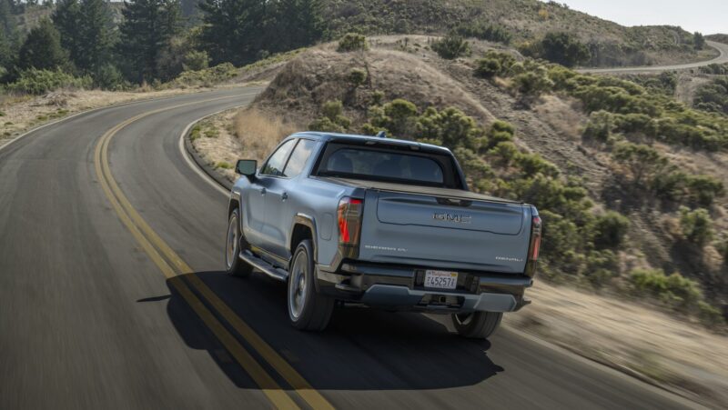 Silver pickup truck driving on a winding road through a hilly landscape with trees and shrubs.