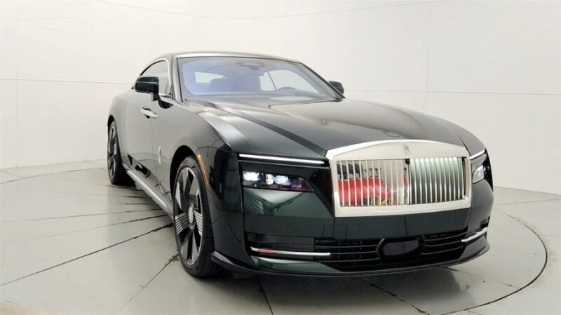 A sleek, dark green Rolls-Royce Spectre graces the showroom with its ultra-luxe design. The electric super coupe boasts a prominent front grille and distinctive headlights, exuding an aura of unmatched elegance and sophistication in the well-lit setting.