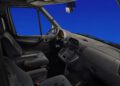 Interior view of a Mercedes-Benz Sprinter's cabin, showcasing the driver's seat, steering wheel, dashboard controls, and passenger seat against a blue background. Celebrating its 30th anniversary, this van blends style and functionality effortlessly.