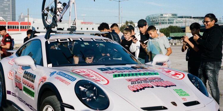 A group of people gather around an exclusive release—a Porsche 911 Dakar—adorned with various decals and a Specialized Bike on the roof, eagerly snapping photos with their phones.
