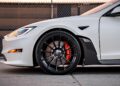 Close-up of a white car's front section, showcasing a sleek black alloy wheel with red brake calipers. The car, reminiscent of the smooth designs popularized by Tesla enthusiasts like Joe Rogan, is parked on a concrete surface near a chain-link fence.