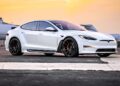 A white Tesla S-APEX sports car is parked on an airport tarmac, with a private jet in the background at sunset, setting the scene for what could be a high-profile delivery or perhaps catching the eye of someone like Joe Rogan.