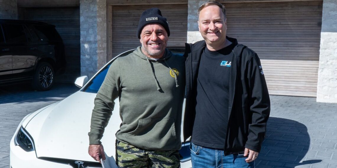 Two men stand smiling in front of a white Tesla S-APEX parked in a driveway. One man wears a beanie and green hoodie, while the other sports a black jacket, clearly pleased with their recent delivery.