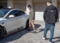 Two people outside near a white Tesla, one wearing a hoodie and shorts, pointing at the vehicle. The other person in jeans and a jacket observes with keen interest.