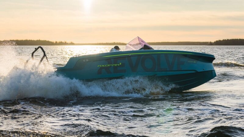 A turquoise speedboat named Revolve creates white waves as it swiftly moves on open water under a sunset sky, offering an exhilarating maritime experience.