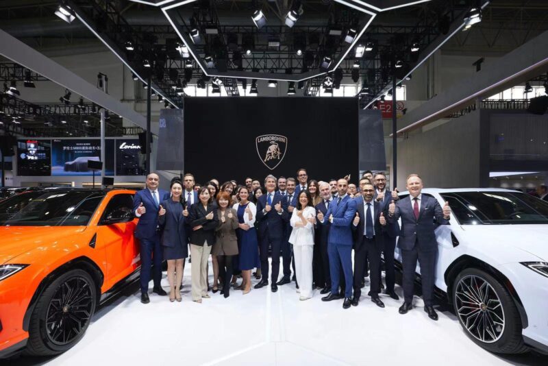 A group of people in business attire stands between two cars in an indoor showroom, celebrating a record-breaking year for Lamborghini car deliveries, as the iconic logo gleams behind them.