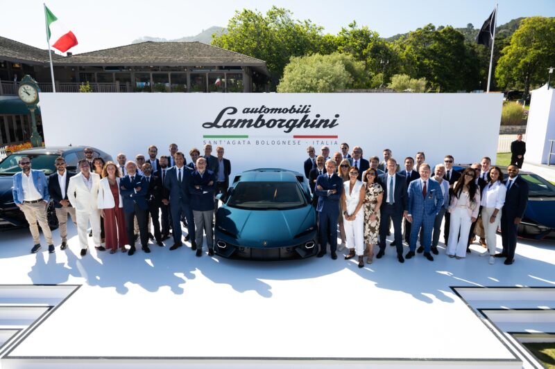 A group of people stands proudly in front of a Lamborghini, celebrating a record-breaking year against a backdrop bearing the Lamborghini logo and "Sant'Agata Bolognese - Italia" text, symbolizing their achievement in car deliveries.