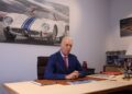 A man in a suit sits at a desk in front of a wall adorned with large car paintings, including the iconic Daytona SP3 emblazoned with historic livery.