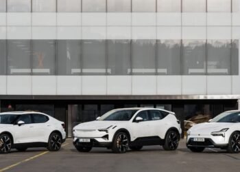 Three white electric cars are parked in front of a modern building with large glass windows.