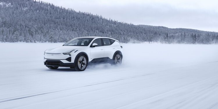A white SUV drives quickly across a snowy landscape with snow-covered trees in the background.