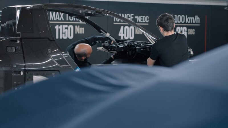 In a workshop adorned with technical specifications, two people meticulously work on assembling a vehicle frame, reminiscent of the craftsmanship seen in Hispano Suiza hypercars.