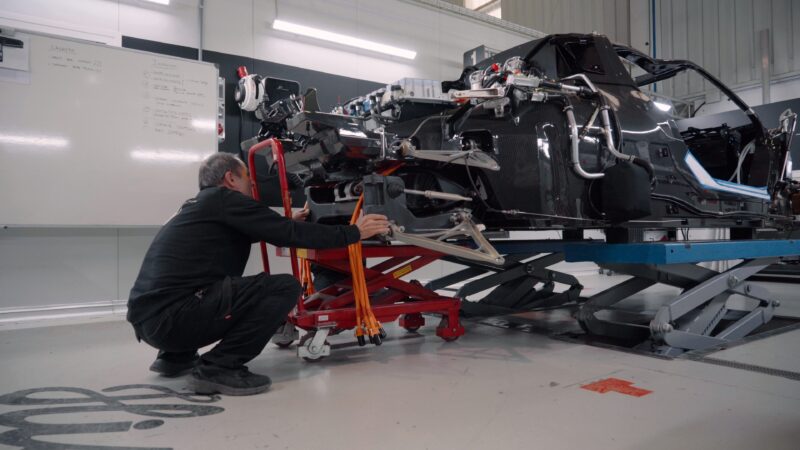 A person kneels beside a partially assembled hypercar chassis in a garage, adjusting components on a hydraulic lift. A whiteboard with notes about the Hispano Suiza Carmen is visible in the background.