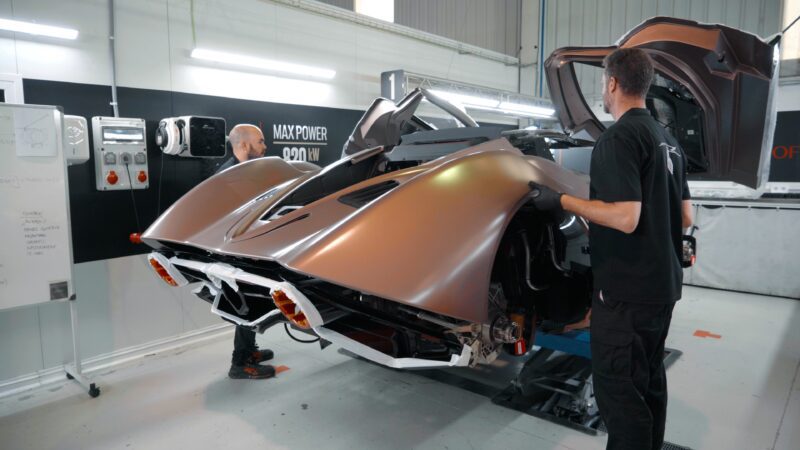 In a workshop, two technicians meticulously work on a futuristic hypercar, its rear compartment wide open. The sleek beauty echoes the craftsmanship of Carmen Sagrera and carries the legacy of Hispano Suiza into a new era.