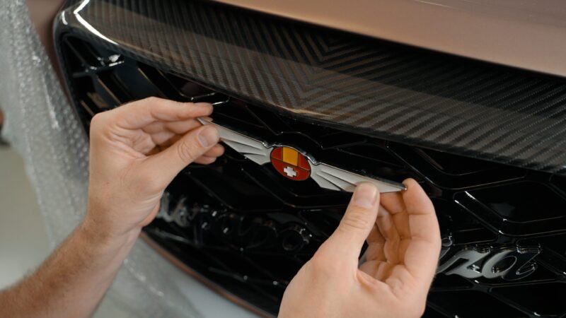 Hands delicately placing the Hispano Suiza emblem on a car grille, showcasing a shield design adorned with a small cross and stripes, reminiscent of the elegance found in hypercars.