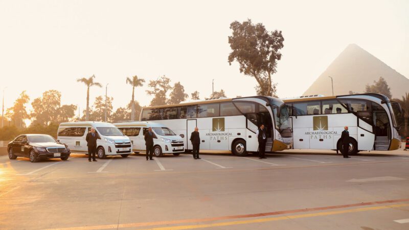 A lineup of luxury travel vehicles, including decorated tour buses and a van, is parked in front of the pyramids. With trees surrounding and several people nearby, this scene sets the stage for an unforgettable experience with Archaeological Paths.